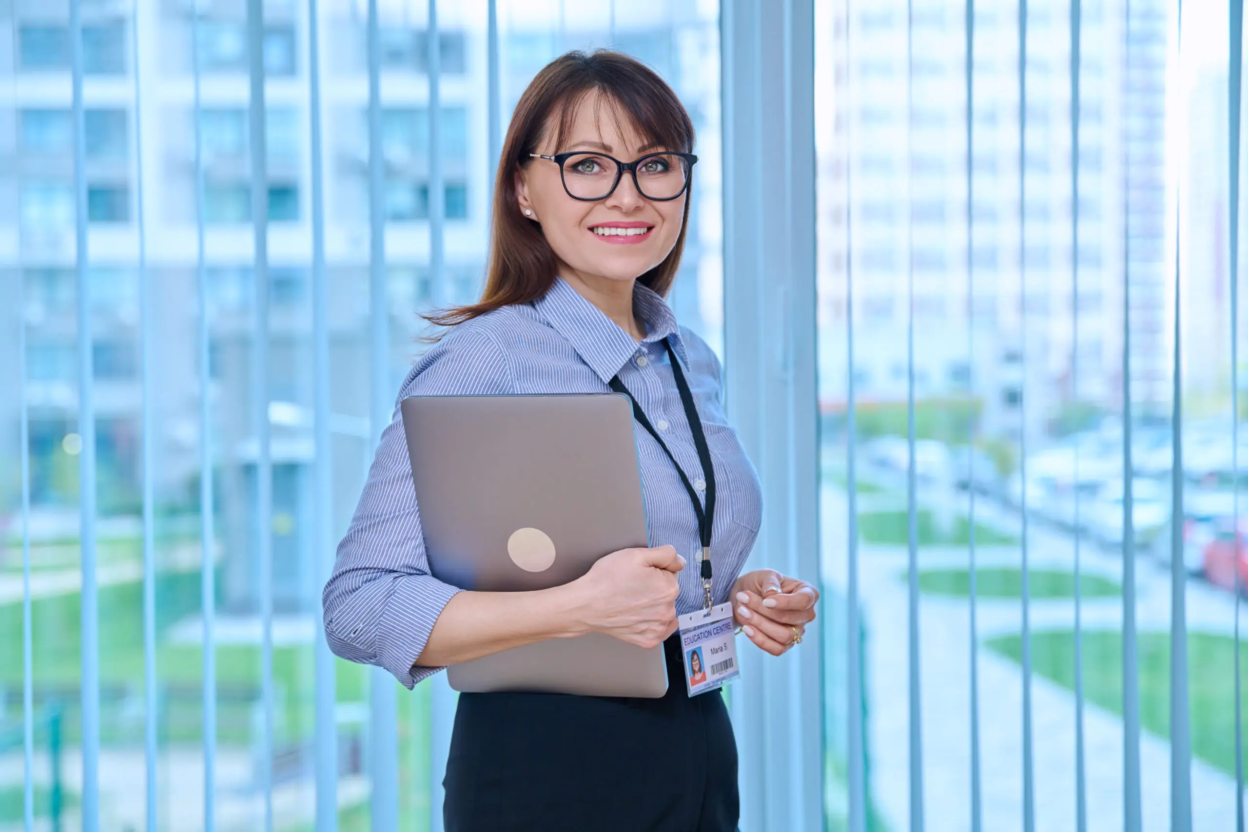 Middle aged woman with educational center id card badge, in office near window A woman holding an open laptop in her hands.